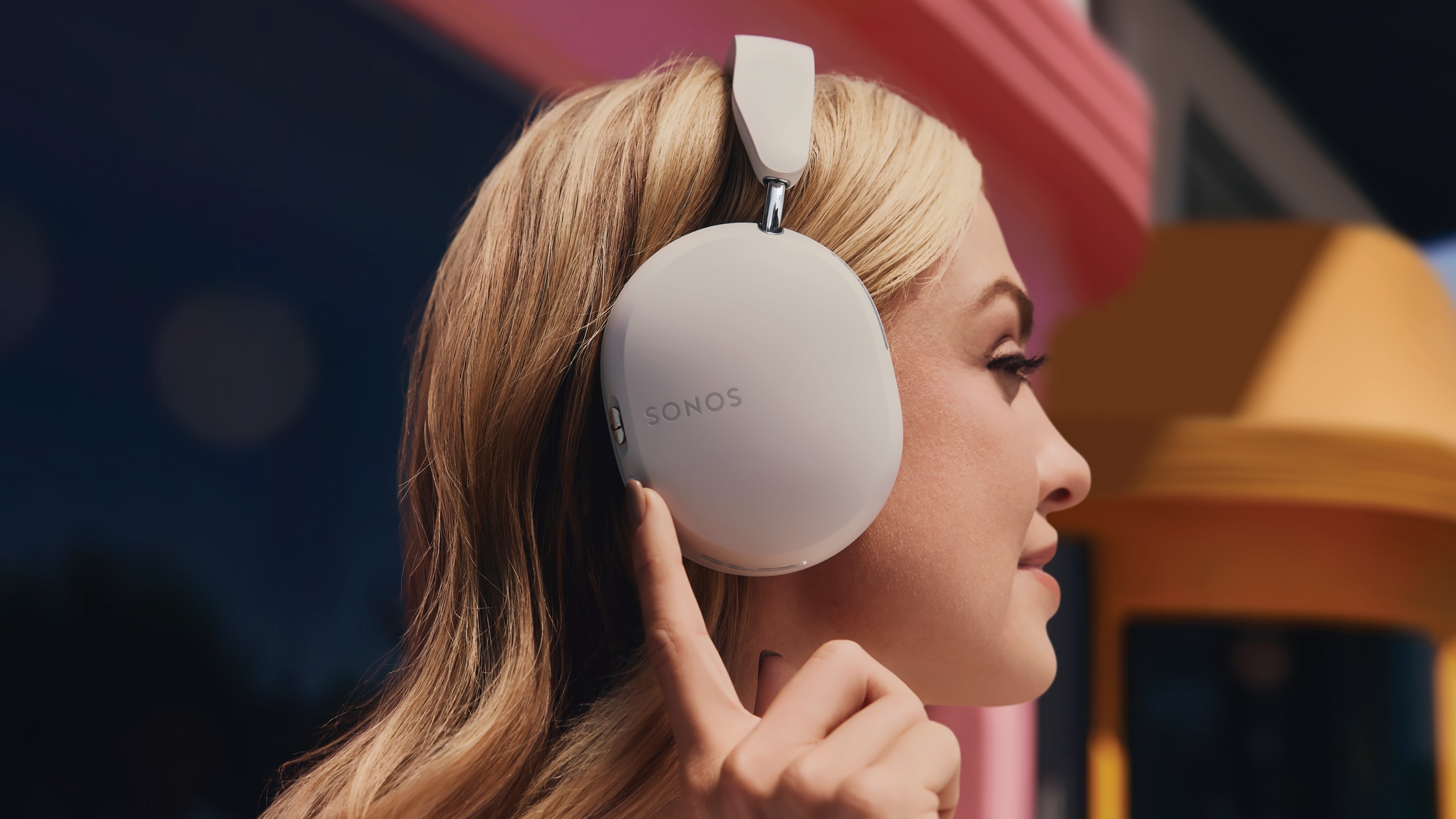 Profilbilde av person som har på et par Soft hvite Sonos Ace-hodetelefoner, og som trykker på knappen for aktiv støydemping 